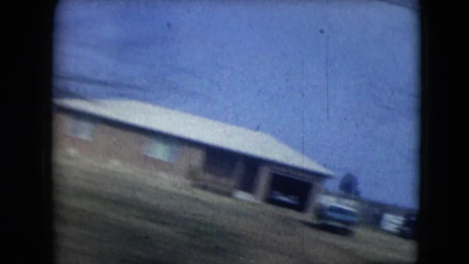 1962 Mexico City home-movie frame showing a blurred low roadside house, open yard, parked car, and dark 8mm frame edges.
