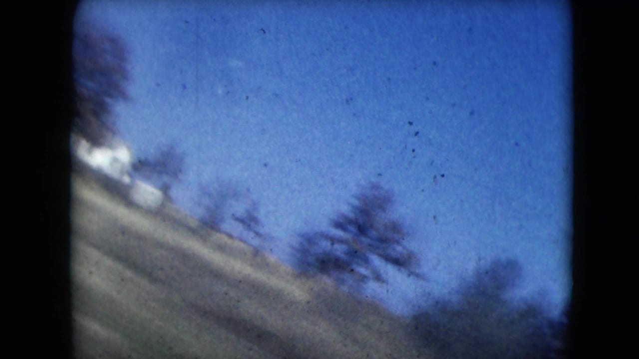 Tilted view of grassy slope, trees, pale structure, and blue sky seen from a moving vehicle.