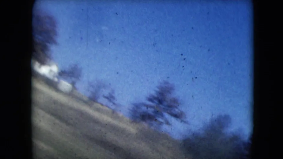1962 Mexico City-area 8mm frame showing a blurred rural roadside slope with trees, a pale distant structure, and blue sky during a fast drive.