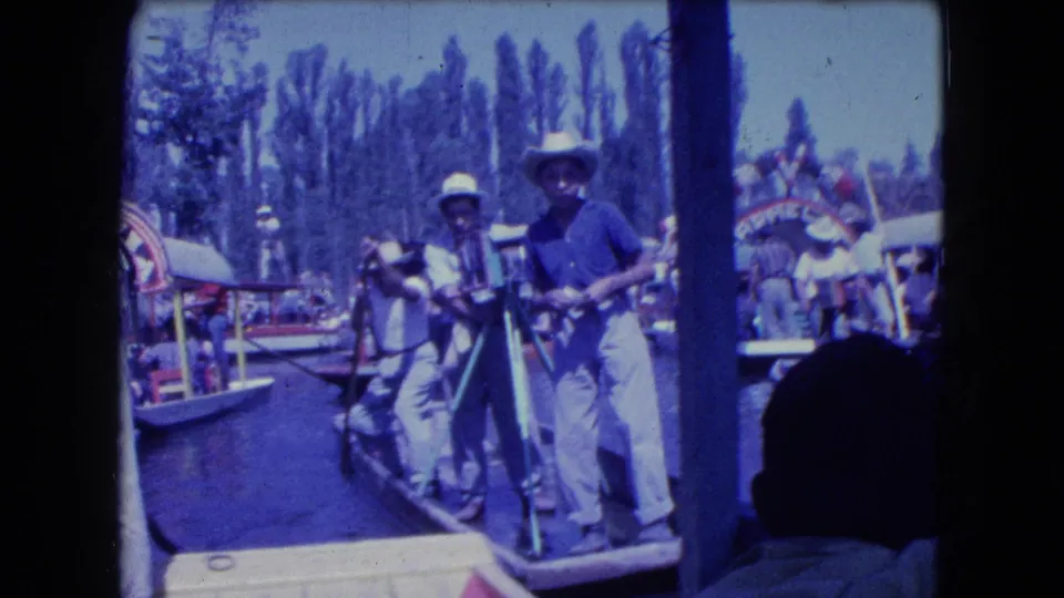 1962 Mexico City canal scene with two boatmen in hats using long poles beside decorated trajineras and passengers.