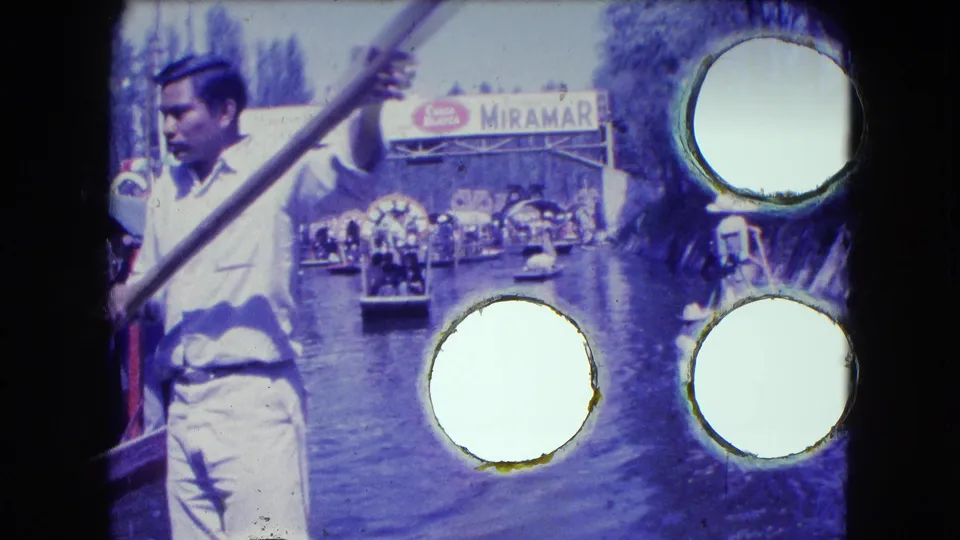 1962 Mexico City canal frame with a boatman, trajineras, passengers, and the MIRAMAR sign partly obscured by film damage.