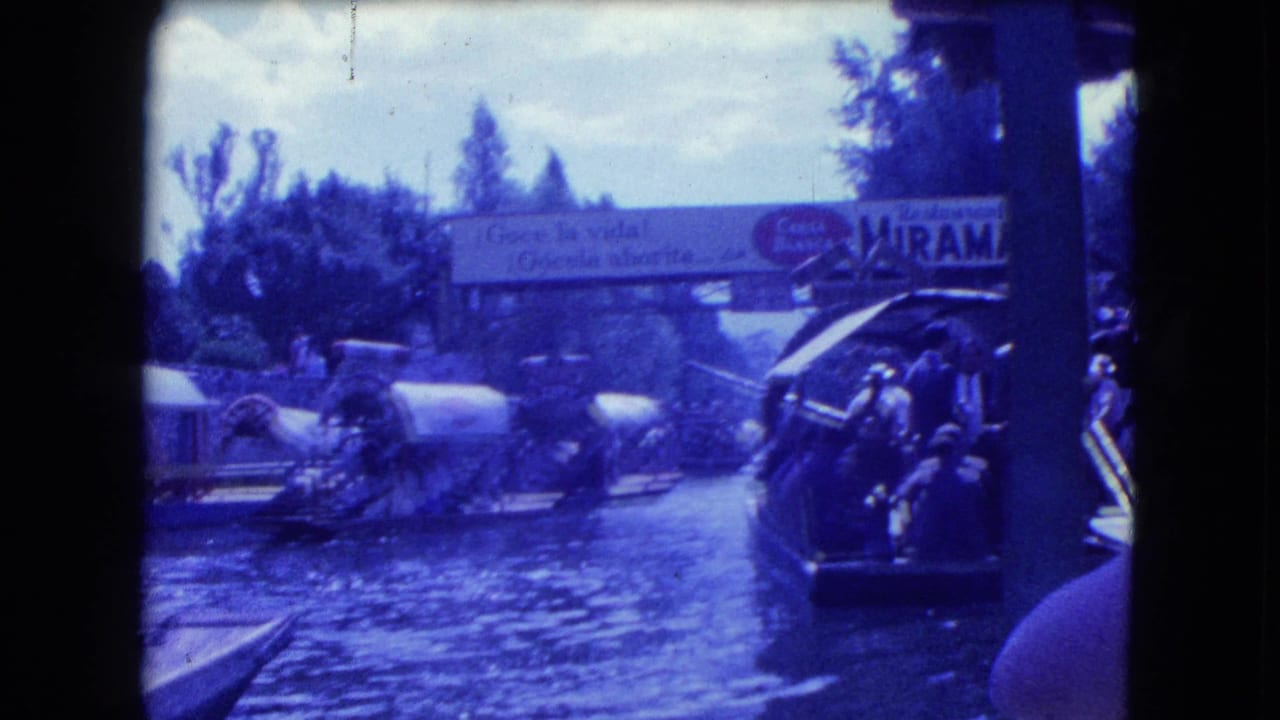 1962 Mexico City home-movie frame showing trajineras on a canal, passengers near the water, trees, and a bridge sign in the background.