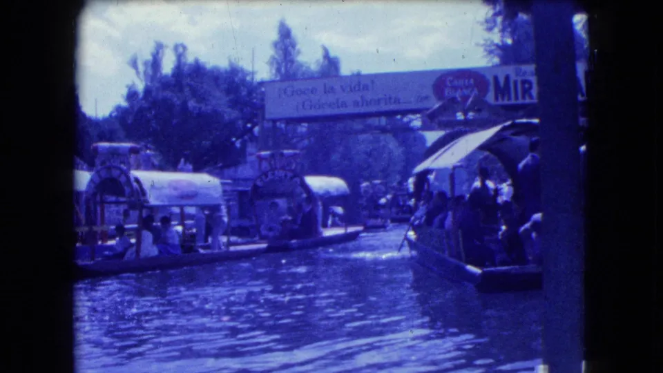 1962 Mexico City home-movie frame showing trajineras and passengers spread across a sunlit canal with trees and signage behind them.