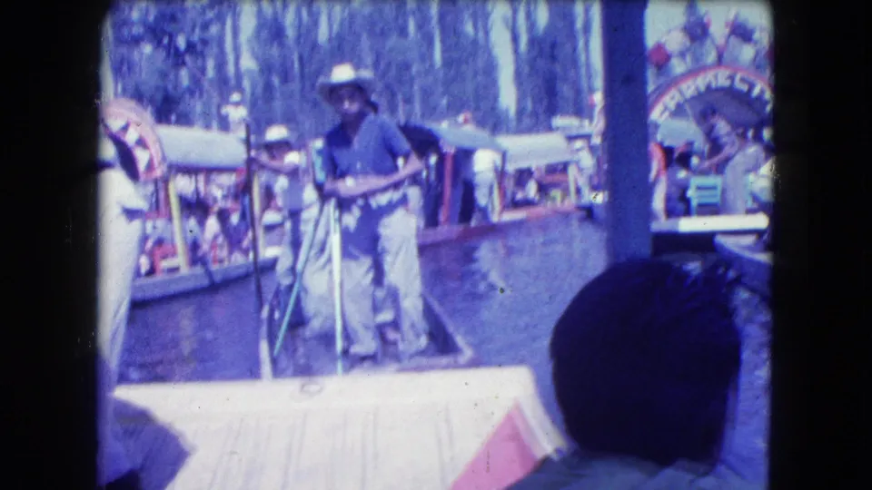 1962 Mexico City canal frame showing a hat-wearing boatman with poles in the foreground and decorated trajineras behind him.