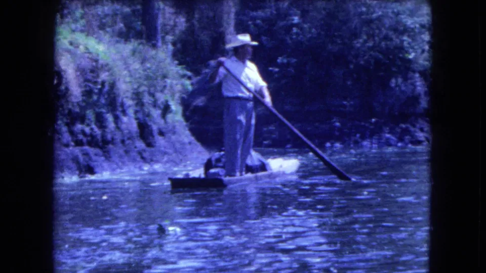 1962 Mexico City canal footage shows a boatman in a straw hat standing on a small boat near a shaded, leafy bank.