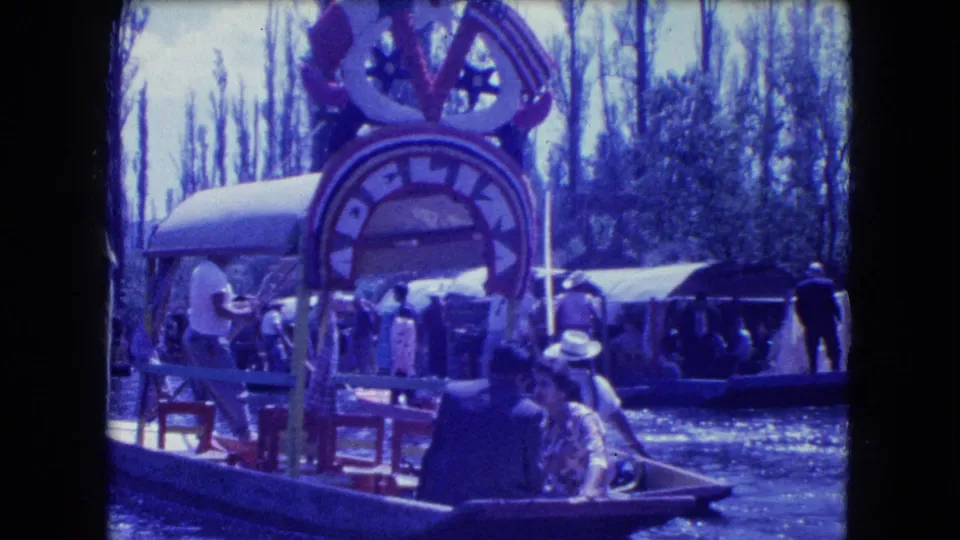 A rower and passengers pass a painted canal arch and other boats in blue-cast 1962 Mexico City home-movie footage.