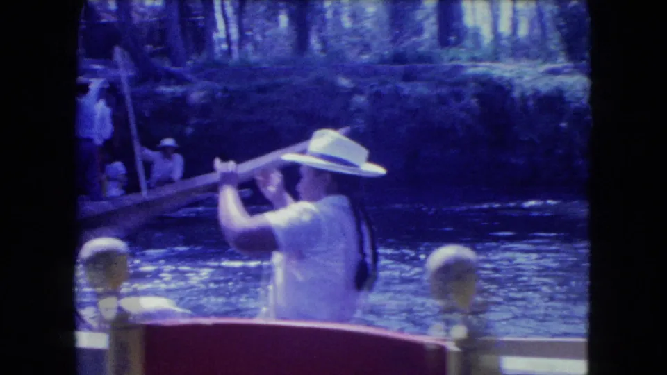 1962 Mexico City frame showing a rower in a wide-brimmed hat using a long oar on the canal, with another boat and people behind.