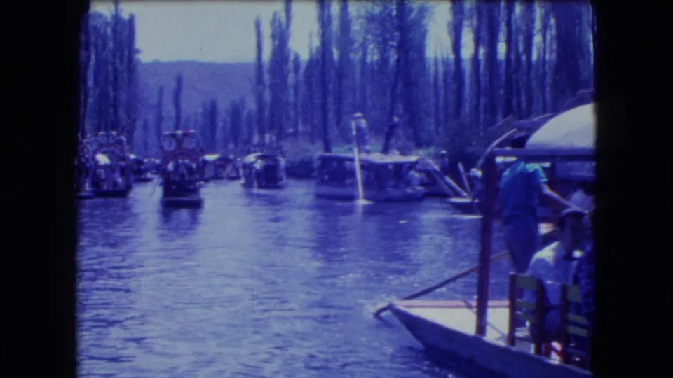 A visible passenger or boat worker stands near a docked craft as trajineras travel on a Mexico City canal.