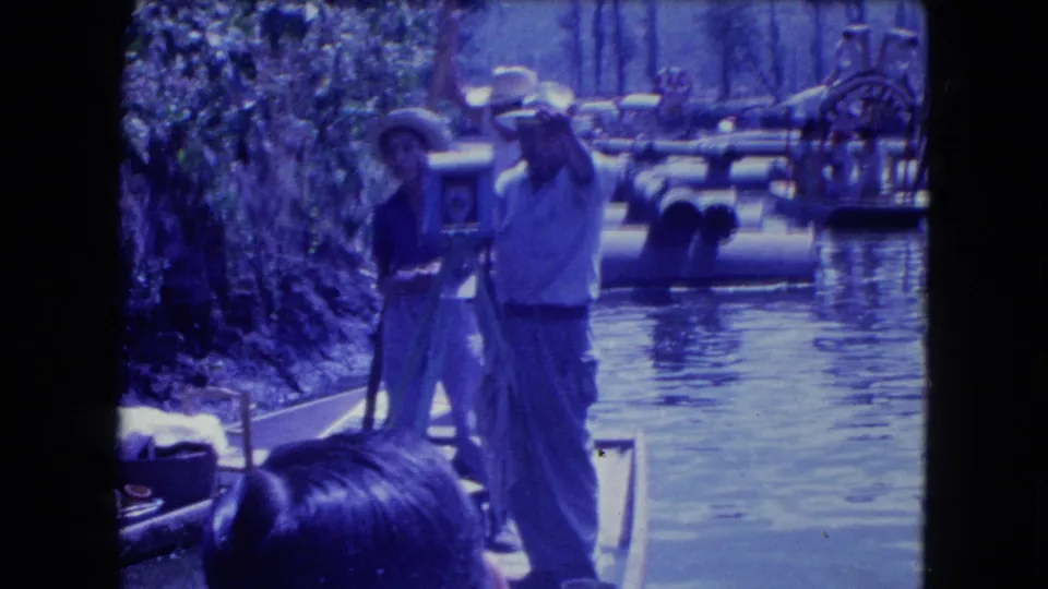 1962 Mexico City canal footage showing a man in a straw hat using a large tripod camera from a boat near other trajineras.