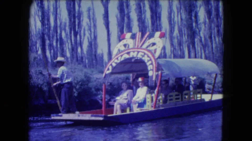 1962 Xochimilco canal scene with a boatman standing at the bow of a decorated trajinera carrying seated passengers.