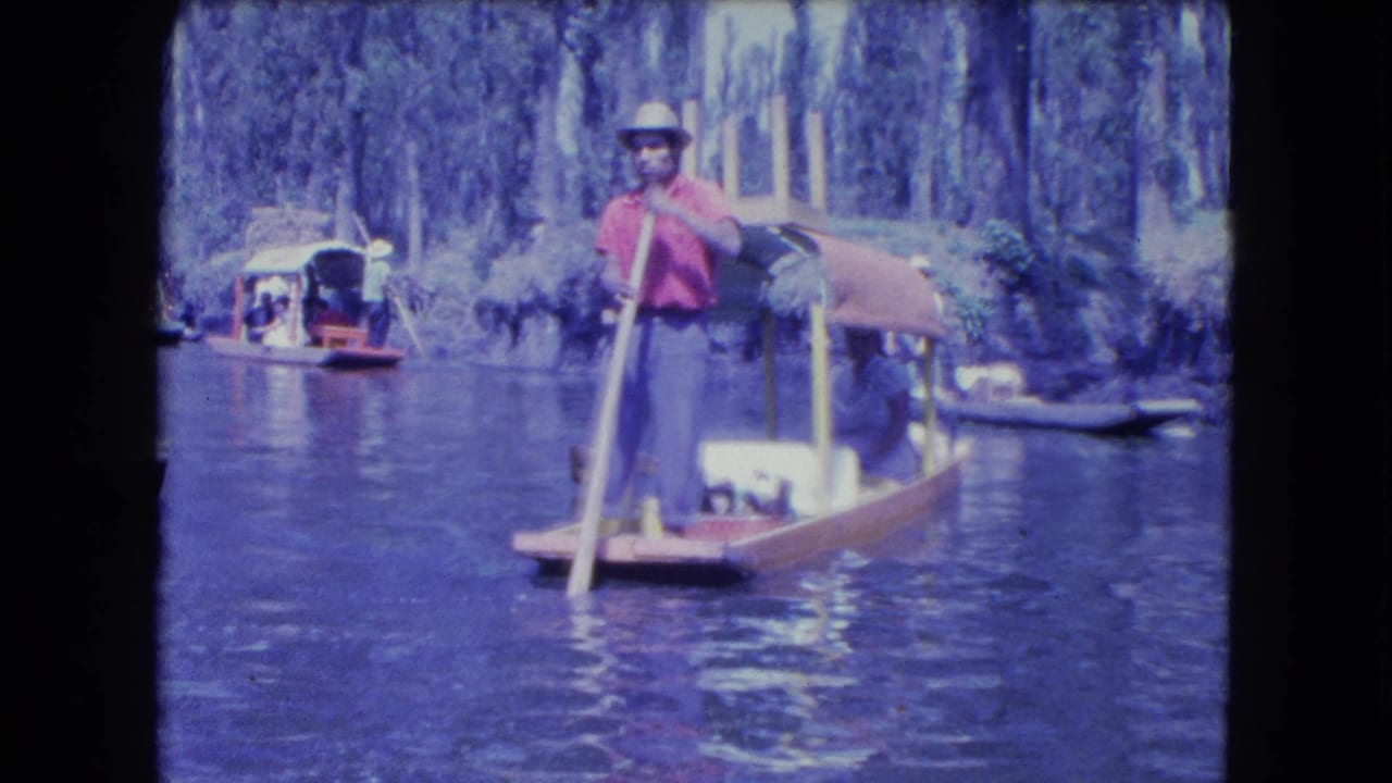 1962 Mexico City canal footage showing a man in a red shirt and hat standing at the stern of a wooden boat with other boats nearby.