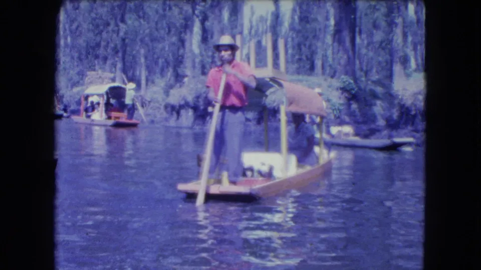 1962 Mexico City canal footage showing a man in a red shirt and hat standing at the stern of a wooden boat with other boats nearby.
