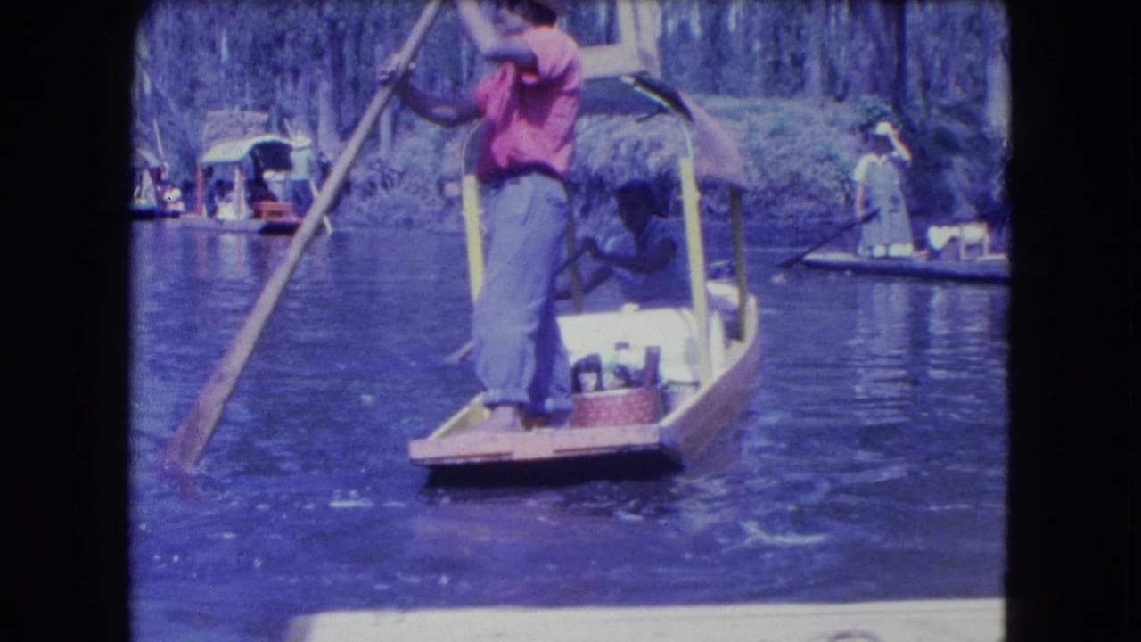 A Mexico City canal boatman in a red shirt pushes a small wooden boat with a pole, with passengers and other boats visible behind him.