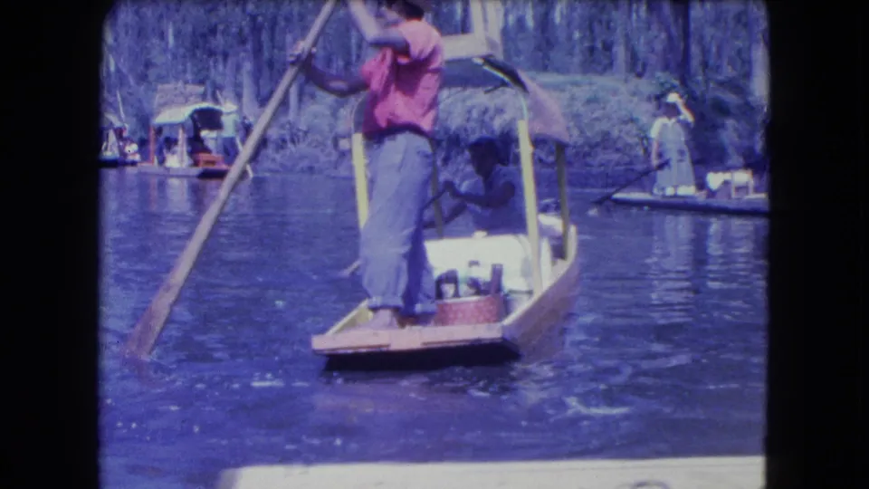 A Mexico City canal boatman in a red shirt pushes a small wooden boat with a pole, with passengers and other boats visible behind him.