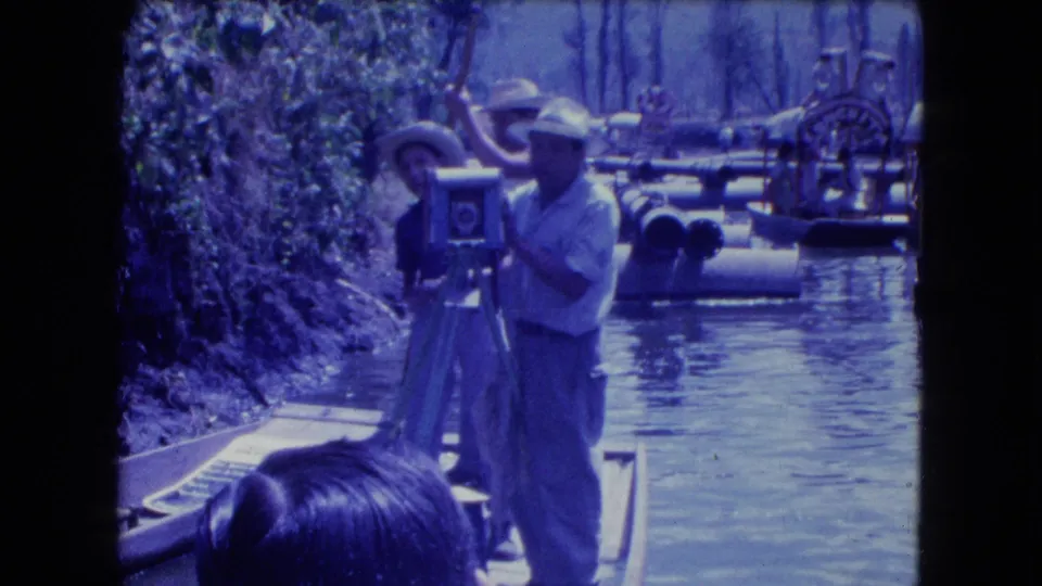 A man stands on a boat beside a large tripod-mounted camera, with canal boats and trees visible behind him in 1962 Mexico City footage.
