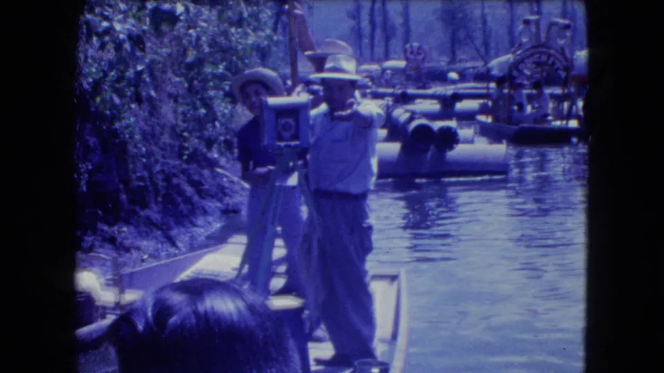 A camera operator on a boat stands next to a large tripod-mounted camera as other canal boats sit or move in the background.