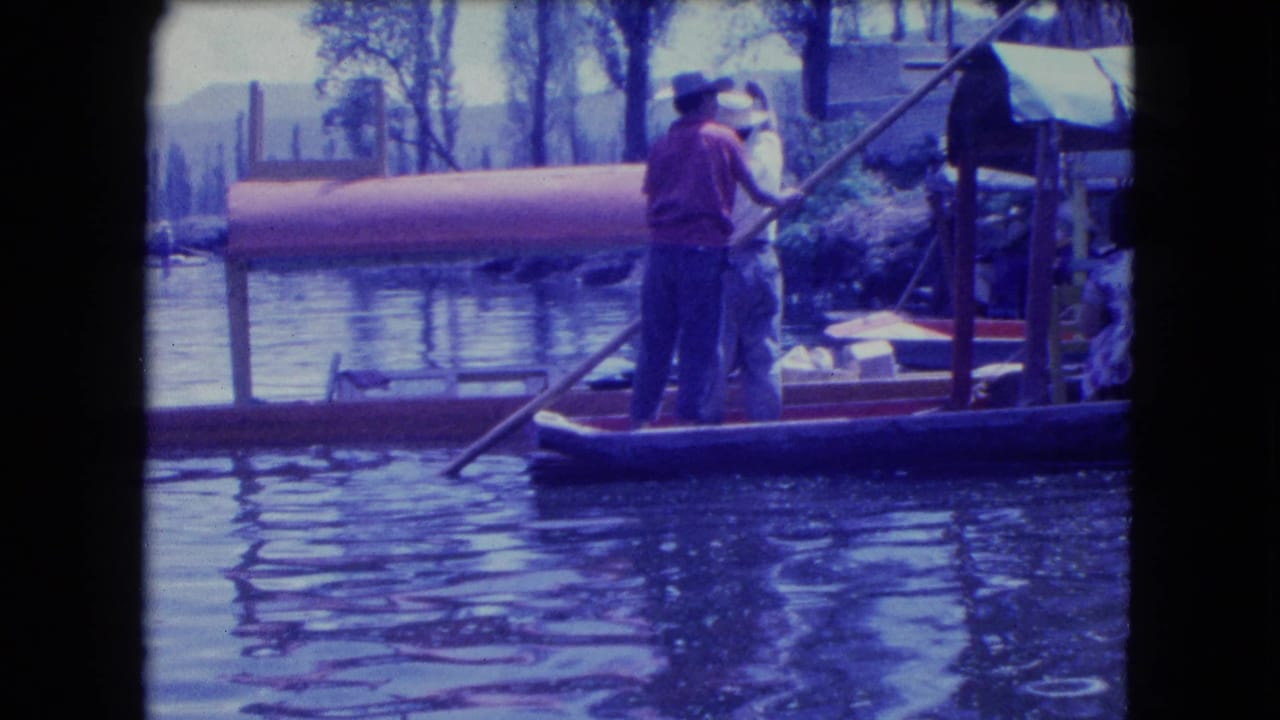 Boat operator uses a long pole on calm water near covered boats.