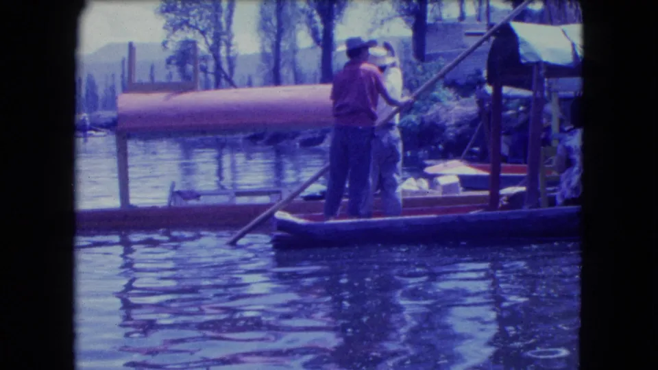 A gondolier-style boat operator in a hat moves a narrow boat with a long pole on a Mexico City waterway.