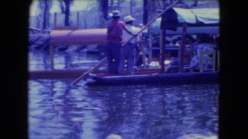 A boat operator stands with a long pole beside covered passenger boats during the 1962 Mexico City excursion footage.