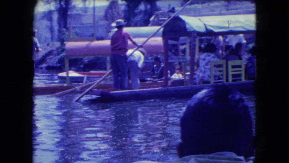 A home-movie view across the water shows a boat operator, covered craft and a foreground spectator in 1962 Mexico City.
