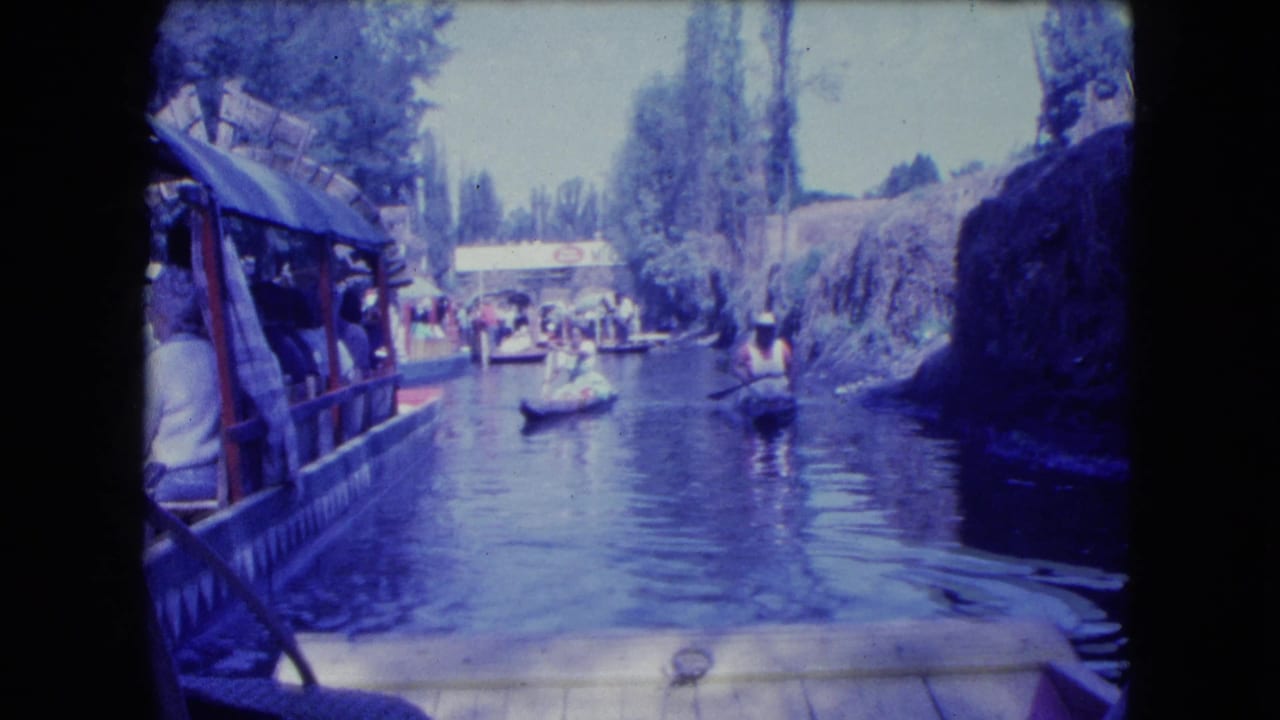 1962 Mexico City canal scene from a moving boat, with a covered passenger boat at left and small paddled craft ahead.