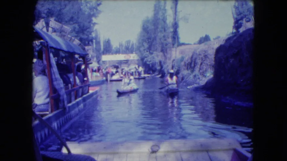 1962 Mexico City canal scene from a moving boat, with a covered passenger boat at left and small paddled craft ahead.
