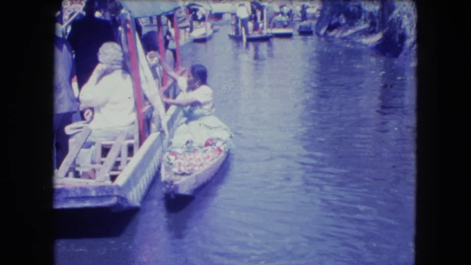 1962 Mexico City canal footage shows a small vendor boat beside a covered passenger boat, with other boats farther ahead.