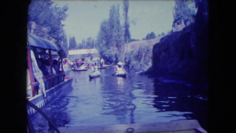 Boat-level 8mm frame of a Mexico City canal with tour boats, distant passengers, tall trees, and reflective water under bright light.