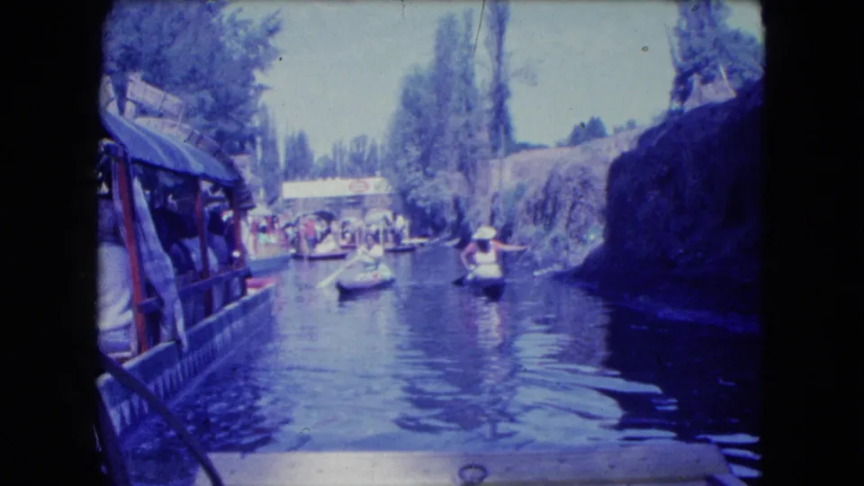 1962 canal-tour frame from Mexico City, showing a small boat on reflective water with a larger covered boat at left and trees beyond.