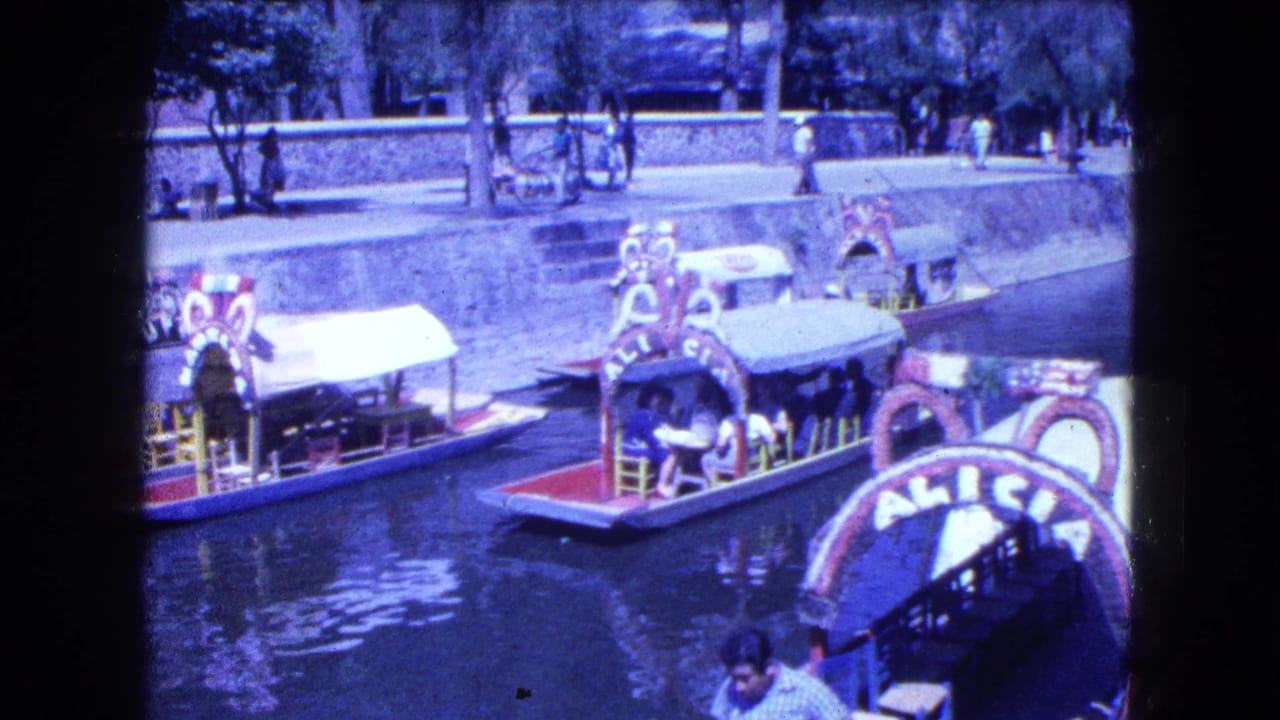 Decorated canal boats on water beside a stone walkway with pedestrians above.