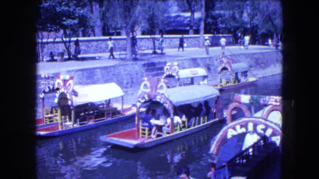 Several colorful canal boats float near a tree-lined walkway with people nearby.