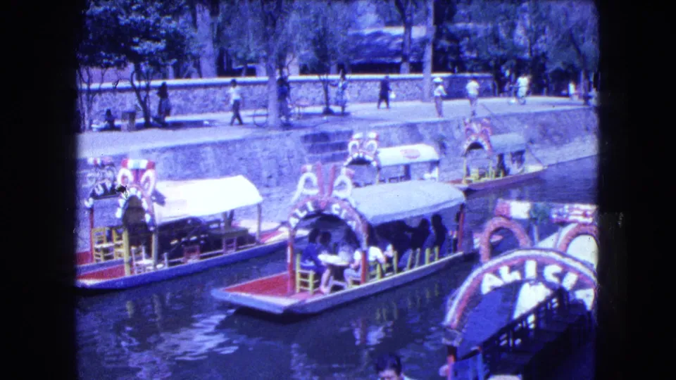 Decorated canal boats in Mexico City appear moored or moving along the canal, with pedestrians on the upper walkway and trees in bright sun.