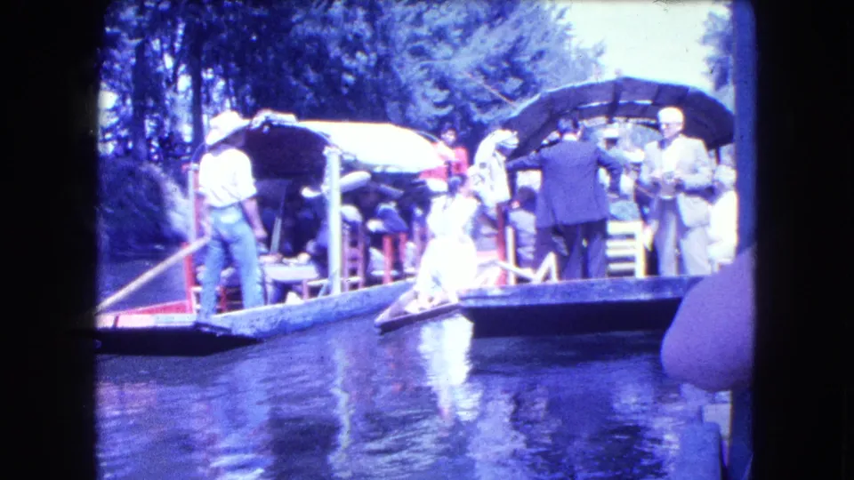 People in period clothing stand and sit on decorated canal boats, with canopies, railings, water reflections, and trees visible behind them.