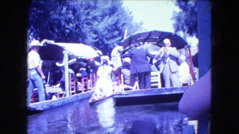 A visitor-shot view shows passengers and attendants on decorated Mexico City canal boats, with bright exposure, trees, and water reflections.