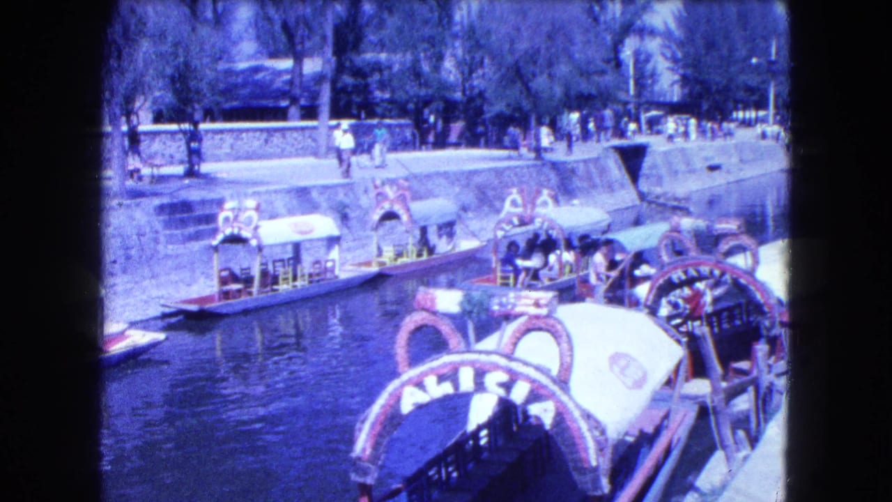 Decorated canal boats with canopies float near a stone embankment as pedestrians walk above.