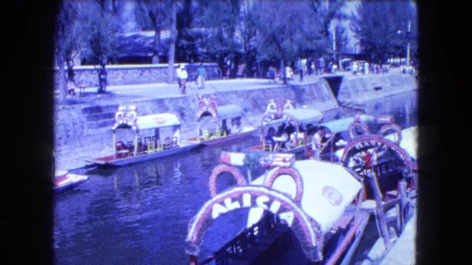 1962 Mexico City canal scene with ornate covered boats, passengers, water, a stone bank, and pedestrians along the walkway.