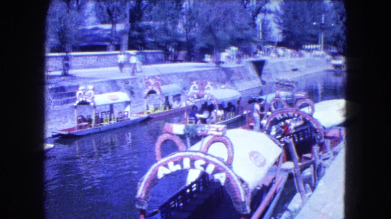 Vintage 8mm view of Mexico City canal boats with ornate canopies, passengers, pedestrians, water, and tree-lined banks.