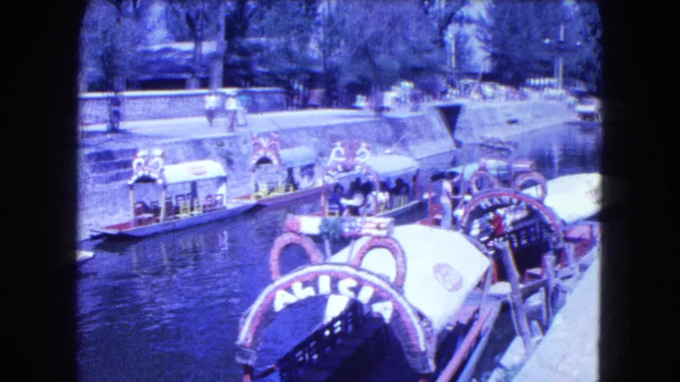 Vintage 8mm view of Mexico City canal boats with ornate canopies, passengers, pedestrians, water, and tree-lined banks.