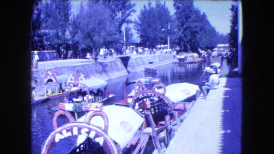 1962 Mexico City home-movie frame showing decorated canal boats near a bright walkway, with passengers and pedestrians in view.