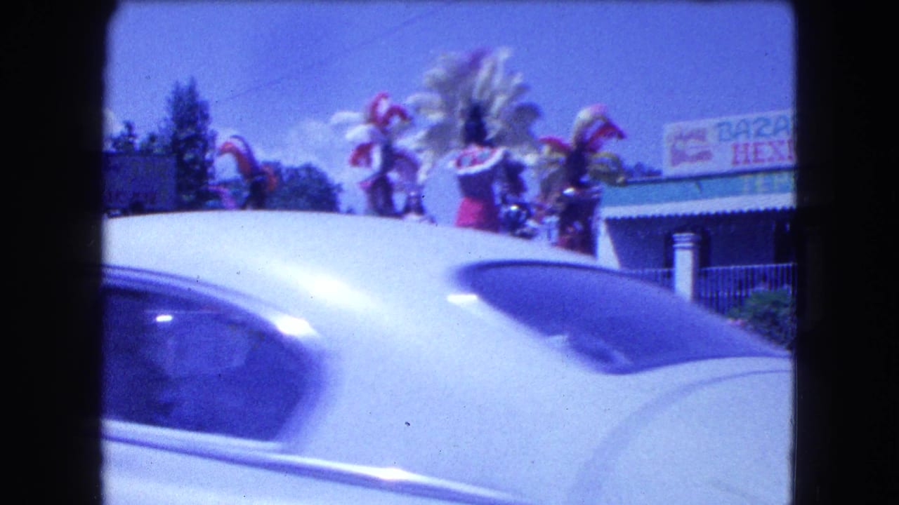 1962 Mexico City home-movie frame with feathered parade performers partly obscured by a passing white car.