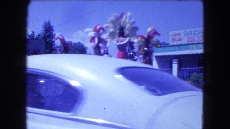 1962 Mexico City home-movie frame with feathered parade performers partly obscured by a passing white car.