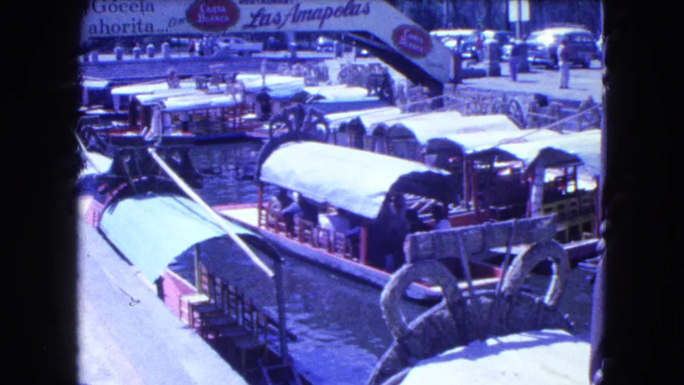 Covered trajineras line the canal near a walkway and large Spanish restaurant sign in 1962 Mexico City footage.