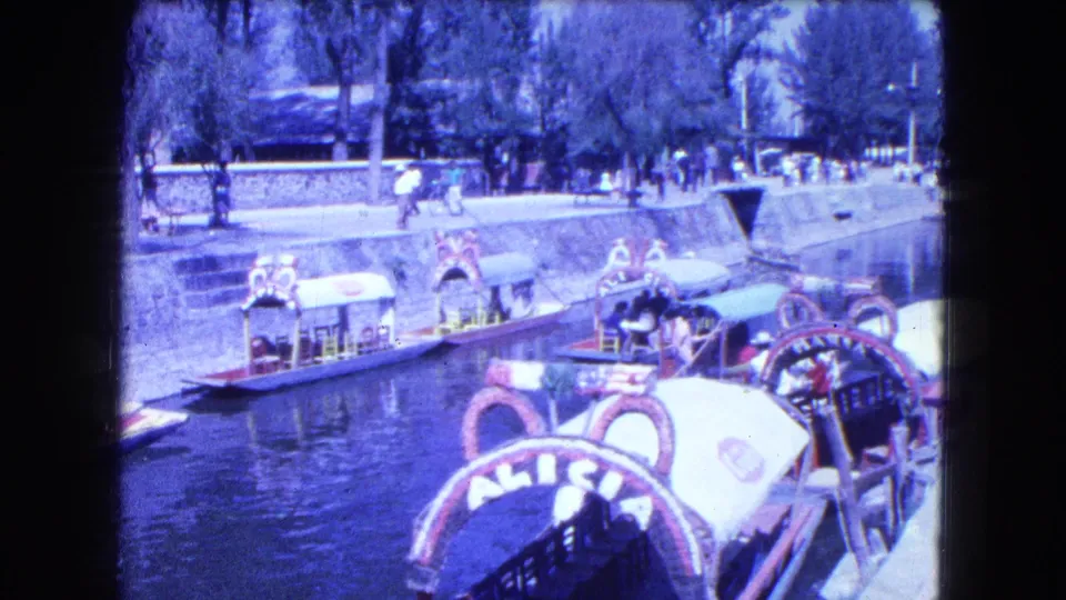 Wider Mexico City canal view with decorated trajineras, pedestrians along the bank, trees, and water traffic.