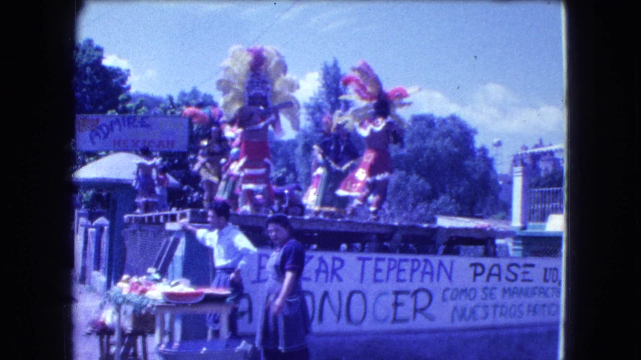 Costumed dancers on a float with a large banner, spectators, vendor goods, and white columns behind them.
