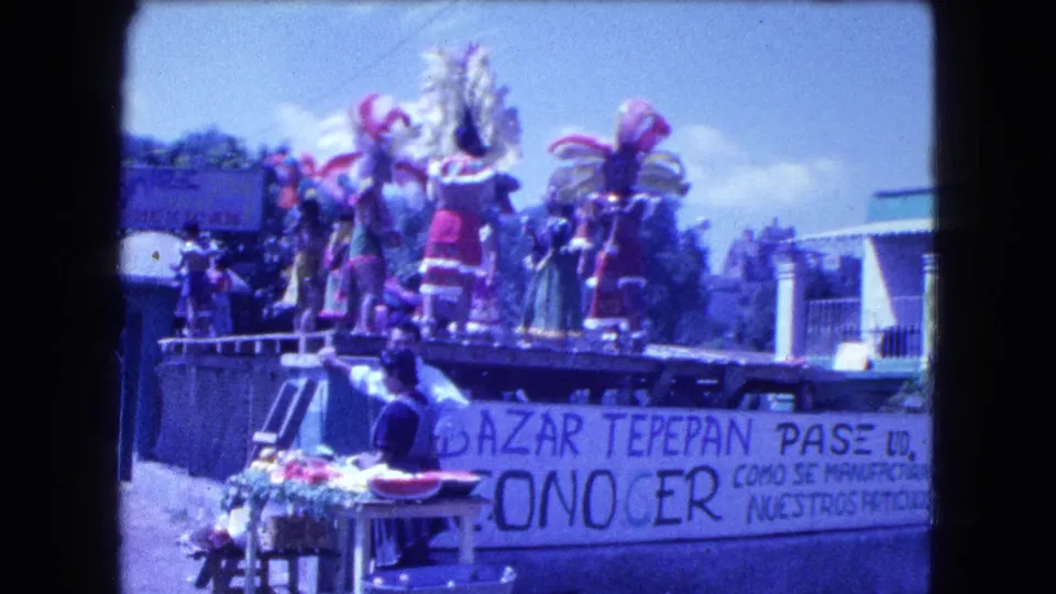 1962 Mexico City home-movie view of feathered dancers on a parade float above a Bazar Tepepan Pase banner and street vendors.