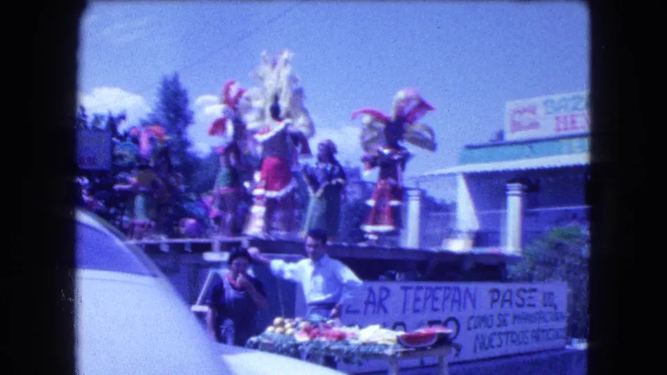 1962 Mexico City street parade frame with costumed dancers on a float, partial Bazar Tepepan Pase banner, spectators, vendors, and a passing car.