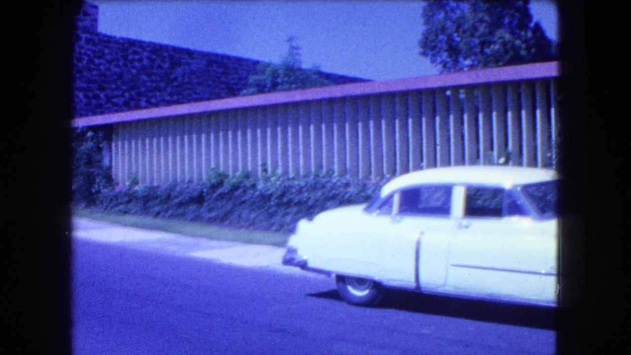 White mid-century car passing a long roadside building on a sunny street.