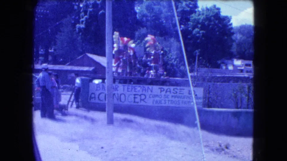 1962 Mexico City home-movie view of bystanders, feathered costumes, a Bazar Tepepan banner, and low roadside buildings.