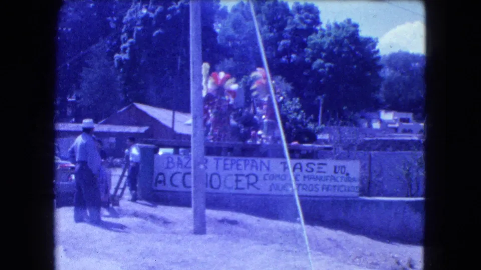 1962 Mexico City street frame showing a man in a hat, a Bazar Tepepan banner, feathered costumes, trees, and low buildings.