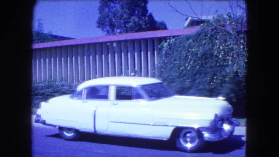 1962 Mexico City home-movie frame with a side view of a white classic sedan, chrome details, curb, hedges, and a residential fence.